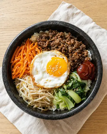 Stone bowl of rice topped with sesame beef mince, a fried egg, carrot, beansprouts, greens and gochujang.