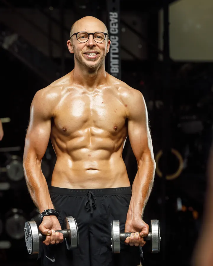 Stuart Rex, founder of Nutrient Warrior, smiling in a gym and holding a pair of dumbbells.